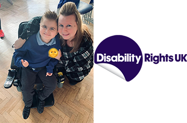 Sally is smiling with her arm wrapped round a young boy in a wheelchair in his school uniform. He is also smiling and is putting his thumb up